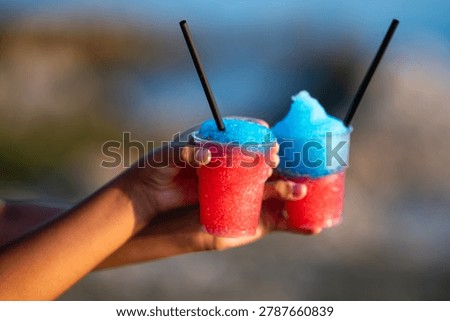 Two hands presenting refreshing red and blue slushy drinks in plastic cups with straws, enjoying summer treats