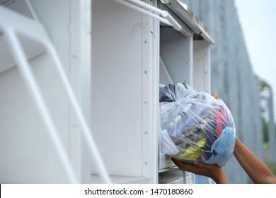 Two Hands Putting A Plastic Bag Full Of Colourful Used Clothing (in Good Condition)  Into An Old White Clothes Donation Bin Or Container For Shoes And Textiles. Recycling And Clothing Bank Outside.