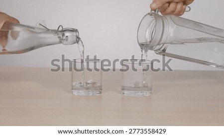 Two hands simultaneously pour fresh water from a glass bottle and pitcher into glasses.