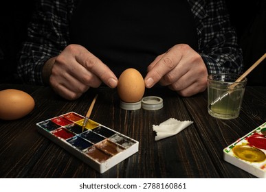 Two hands work on decorating an egg with paint during a craft session. A paint palette and a jar sit nearby. The setting is a simple wooden table in a dimly lit room.