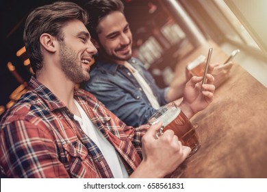 Two handsome male friends in bar drinking beer with smartphones in hands and smiling. - Powered by Shutterstock