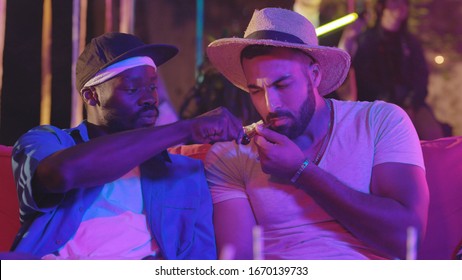 Two Handsome Male Friends Resting At Summer Party. Attractive Caucasian Guy Trying Weed Pipe Coughing Of Smoke. Illegal Party Entertainment.