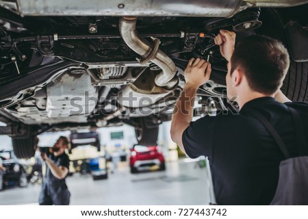 Two handsome mechanics in uniform are working in auto service with lifted vehicle. Car repair and maintenance.