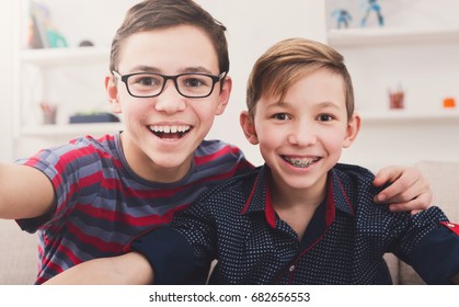 Two handsome teenage boys taking selfie while posing with smile to phone at home. Wearing glasses and braces. Friendship, brotherhood and health care concept