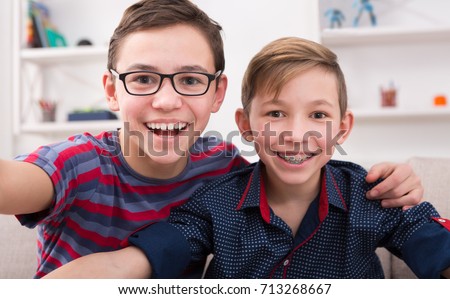 Two handsome teenage boys taking selfie while posing with smile to phone at home. Wearing glasses and braces. Friendship, brotherhood and health care concept