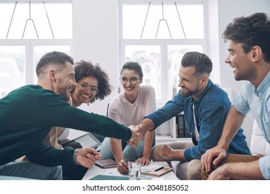 Two Handsome Young Men Shaking Hands And Smiling While Having Meeting With Colleagues In Office