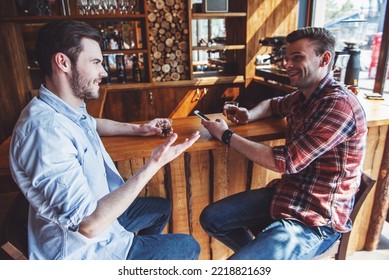 Two handsome young men are using a smartphone, drinking, talking and smiling while sitting at bar counter in a modern urban cafe