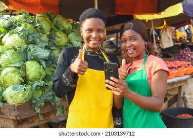 Two happy African business women or female traders wearing colorful aprons while standing at a vegetable stall in a market place with a smart phone with them