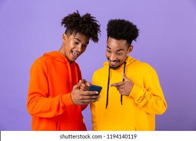 Two Happy African Man Friends Dressed In Colorful Hoodies Standing Isolated Over Violet Background, Looking At Mobile Phone