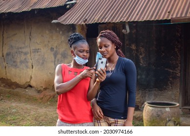 Two happy african women or ladies wearing nose mask, stands outside a village mud house, holding and looking into a smart phone with them