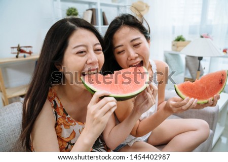 two happy asian chinese female friends eating watermelon sharing one slice together sitting on comfort couch in living room with air conditioner electric fan wind breeze blowing hair indoors summer