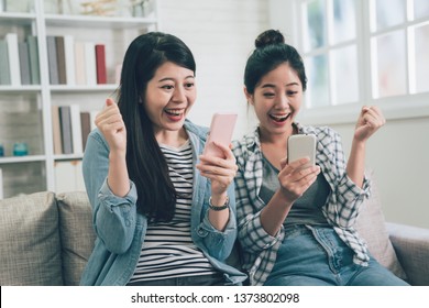 Two Happy Asian Female Friends Sitting On Couch Sofa At Home In Living Room Smiling Laughing With Cheerful Face Win Hands Gesture Staring At Smart Phone. Young Women Using Cellphone Indoors Joyful.