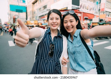 two happy asian tourist girls are smiling at the camera with thumb up gesture while taking a selfie on the street of a sightseeing shopping area in summer.