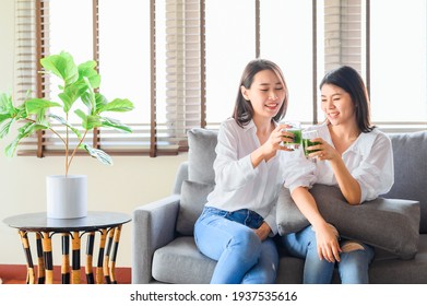 Two Happy Asian Woman Friends Enjoy Drinking Green Juice Together In Living Room