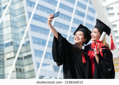 Two Happy Asian Young Beautiful Graduate Female Students With University Degree Standing And Holding Diploma Taking Selfie Picture With Mobile Phone After Graduation. Blur Background Of Building