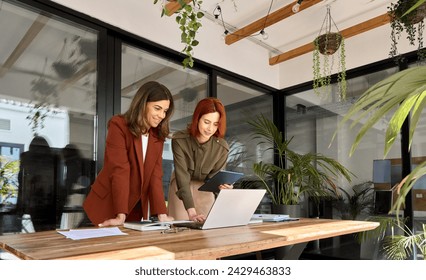 Two happy business women executives of young and middle age talking working in green office. Smiling professional ladies partners using laptop and tablet technology devices standing at work table. - Powered by Shutterstock