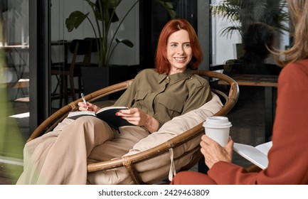 Two happy business women talking in cozy office sitting in chairs. Smiling professional ladies colleagues partners having conversation drinking coffee having conversation taking notes at work. - Powered by Shutterstock