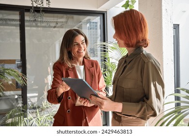 Two happy business women of young and middle age talking standing in creative green office space. Smiling professional ladies employees colleagues having conversation using digital tablet at work.