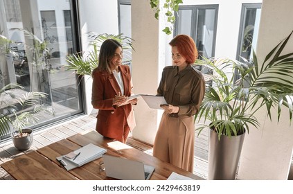 Two happy business women of young and middle age talking in creative green office space. Smiling professional ladies entrepreneurs workers partners having conversation using technology devices at work - Powered by Shutterstock