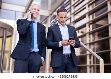 Two happy businessmen standing outside office building talking on a smartphone - Powered by Shutterstock