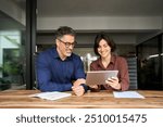 Two happy busy business people executives team talking sitting in office. Busy mature professional business man and woman partners working together having conversation in office using digital tablet.