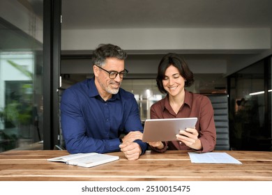 Two happy busy business people executives team talking sitting in office. Busy mature professional business man and woman partners working together having conversation in office using digital tablet. - Powered by Shutterstock