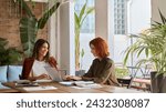 Two happy busy business women of young and middle age talking in green cozy office sitting at desk. Professional ladies executives having conversation using laptop at work. Authentic candid shot.