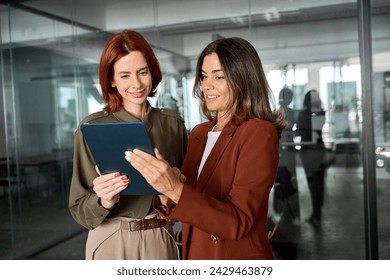 Two happy busy business women talking using tab working together standing in office. Professional female colleagues employees looking at digital tablet tech discussing corporate project at work. - Powered by Shutterstock