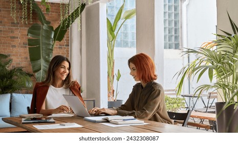 Two happy busy business women of young and middle age talking in green cozy office sitting at desk. Professional ladies executives having conversation using laptop at work. Authentic candid shot. - Powered by Shutterstock