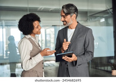 Two happy busy diverse professional business team people talking standing in office using tab. Smiling young African woman employee and older Latin man executive having conversation at work. - Powered by Shutterstock