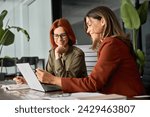 Two happy busy female employees working together using computer planning project. Middle aged professional business woman consulting teaching young employee looking at laptop sitting at desk in office