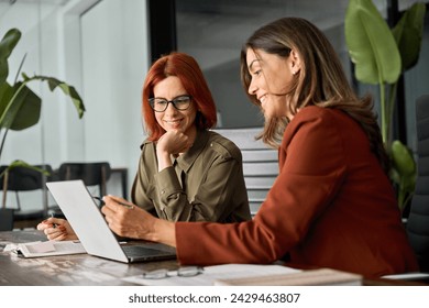 Two happy busy female employees working together using computer planning project. Middle aged professional business woman consulting teaching young employee looking at laptop sitting at desk in office - Powered by Shutterstock