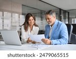 Two happy busy middle aged professionals man and woman business leaders partners checking document reading financial report talking working together on laptop computer in office at corporate meeting.