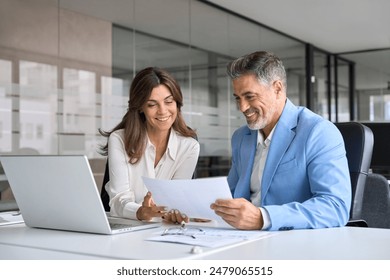 Two happy busy middle aged professionals man and woman business leaders partners checking document reading financial report talking working together on laptop computer in office at corporate meeting.