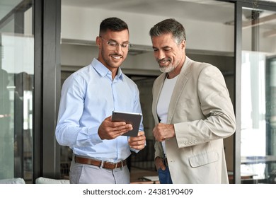 Two happy busy professional business men executive business team working together standing in office holding tab using digital tablet computer tech managing data standing in corporate office. - Powered by Shutterstock