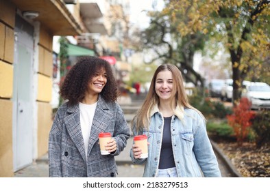 Two Happy Carefree Multiracial Girlfriends Walking City Street With Take Away Coffee, Talking And Sharing Life Stories, Multiethnic Female Best Friends Enjoying Autumn Walk Outdoors. Womens Friendship