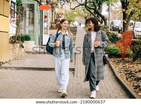 Two happy carefree multiracial girlfriends walking city street with take away coffee, talking and sharing life stories, multiethnic female best friends enjoying autumn walk outdoors. Womens friendship