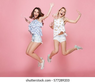 Two happy carefree young women jumping and listening to music from cell phones over pink background