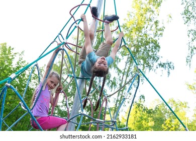 Two happy caucasian girls having fun on playground, climbing the rope net. The older girl is hanging upside down