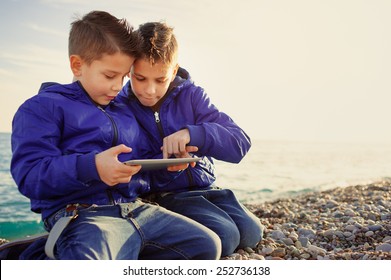 Two Happy Caucasian Kids, Brothers, Playing Together With Tablet Pc Sitting Outdoors At Pebble Beach Against The Sea