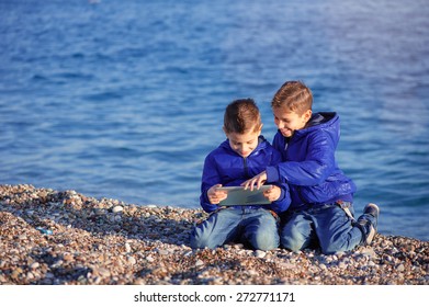 Two Happy Caucasian Kids, Brothers, Playing Together With Tablet Pc Sitting Outdoors At Pebble Beach Against The Sea