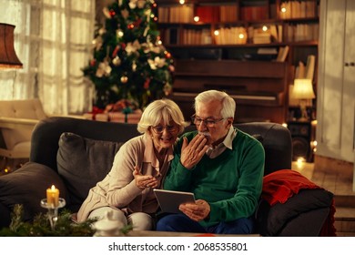 Two happy and cheerful senior sitting on the sofa using together the same tablet or technology device the christmas day - senior people having fun and sending kiss to family - Powered by Shutterstock