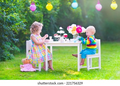 Two happy children, cute curly toddler girl and a little baby boy, brother and sister, enjoying a tea party with their toys playing with dishes, cup cakes and muffins in a sunny summer garden