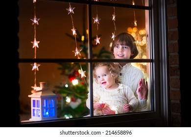 Two Happy Children, Little Curly Toddler Girl And Laughing Boy In Warm Knitted Winter Sweaters Standing Next To A Window In A Decorated Living Room With Christmas Tree, View From Outside Of The House