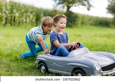 Two Happy Children Playing With Big Old Toy Car In Summer Garden, Outdoors. Boy Driving Car With Little Girl Inside. Laughing And Smiling Kids. Family, Childhood, Lifestyle Concept.