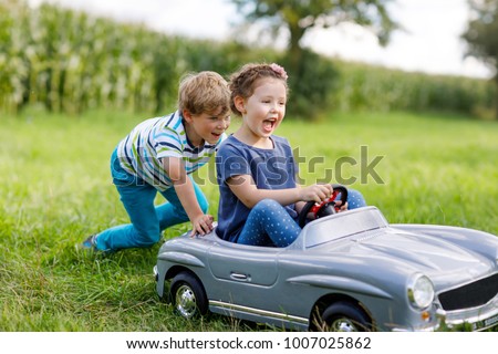 Two happy children playing with big old toy car in summer garden, outdoors. Boy driving car with little girl inside. Laughing and smiling kids. Family, childhood, lifestyle concept.