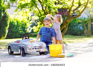 Two happy children washing big old toy car in summer garden, outdoors. Brother boy and little sister toddler girl cleaning car with soap and water, having fun with splashing and playing with sponge. - Powered by Shutterstock