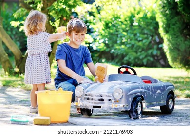 Two Happy Children Washing Big Old Toy Car In Summer Garden, Outdoors. Brother Boy And Little Sister Toddler Girl Cleaning Car With Soap And Water, Having Fun With Splashing And Playing With Sponge.