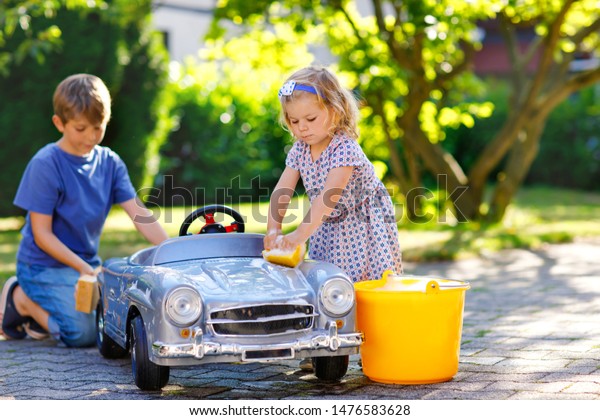 Two happy children washing big old toy car in\
summer garden, outdoors. Brother boy and little sister toddler girl\
cleaning car with soap and water, having fun with splashing and\
playing with sponge.