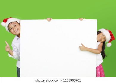Two Happy Children Wearing Santa Hat While Showing Thumbs Up And Blank Whiteboard. Shot With Green Screen